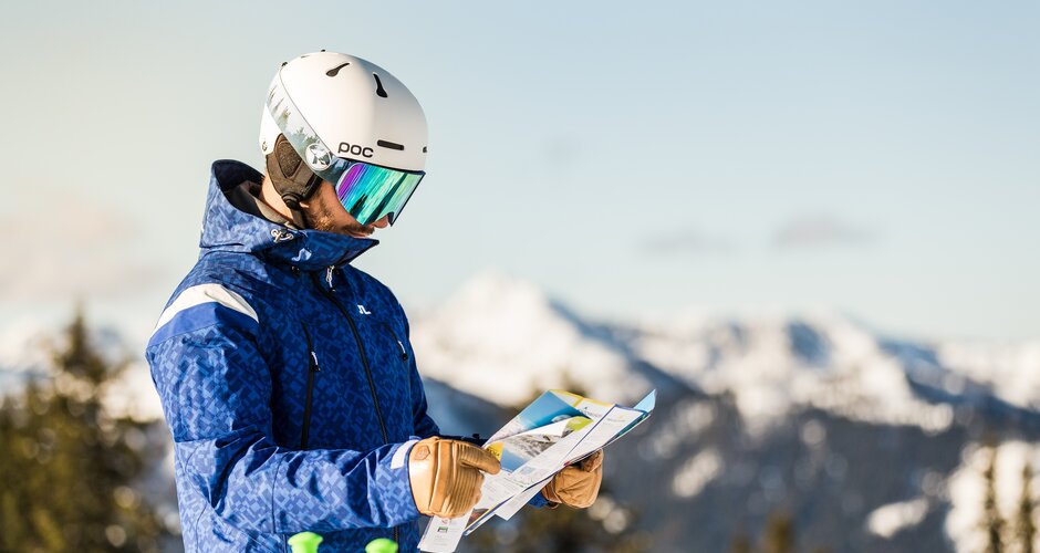 Skifahrer in blauer Jacke und weißem Helm schaut auf Pistenplan vor Alpenpanorama. | © Dominik Steiner