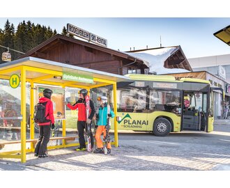 Skifahrer mit Ausrüstung an der Skibusstation Hochwurzen, Skibus im Hintergrund. | © David Stocker