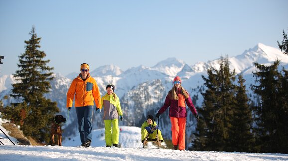 Familie mit Schlitten und Hund wandert auf verschneitem Weg mit Alpenpanorama. | © Gregor Hartl