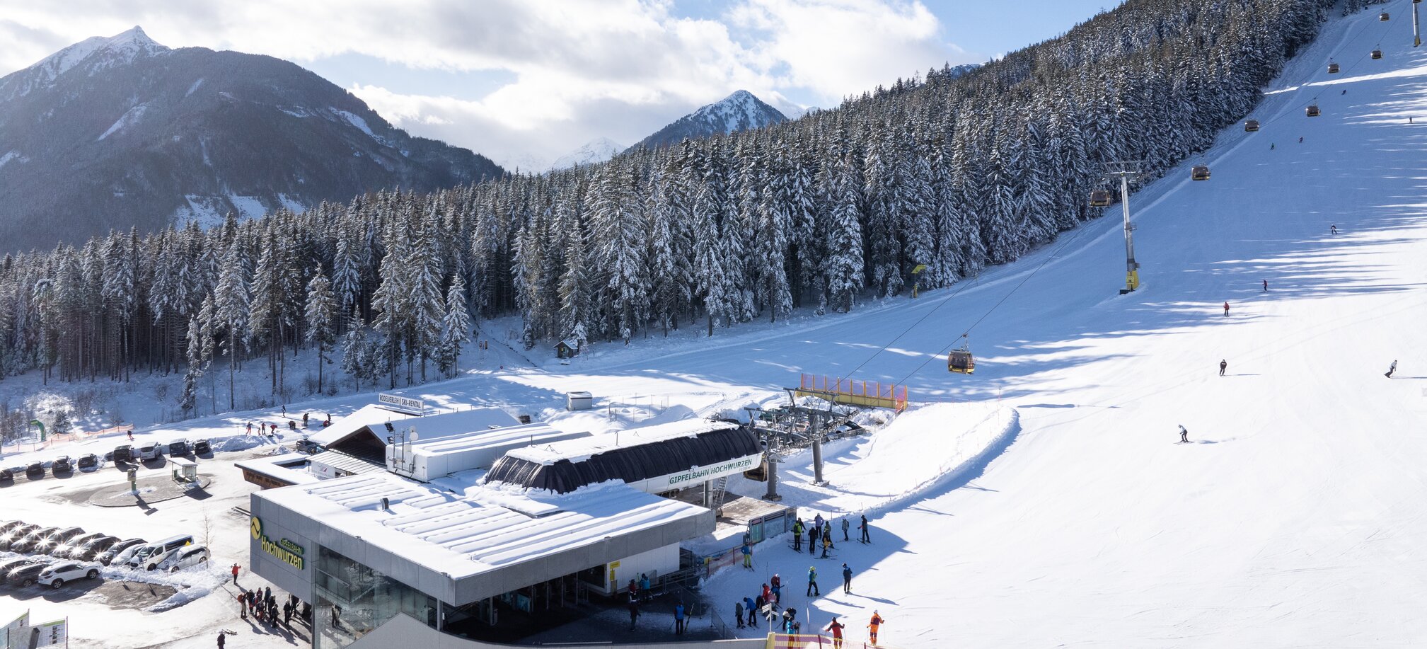 Blick auf die Talstation, Skifahrer und Gondeln, Wald und Berge im Hintergrund. | © Josh Absenger