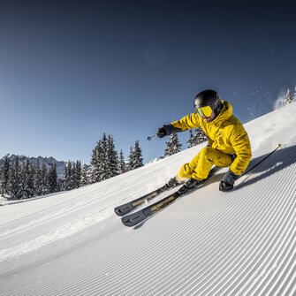 A skier in yellow outfit speeds down a freshly groomed slope, carving turns as snow sprays into the air. | © Mirja Geh