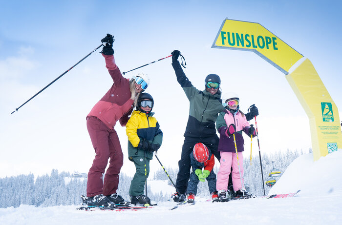 Glückliche Familie auf Skiern und mit Skianzug steht beim Start einer Funslope | © Josh Absenger