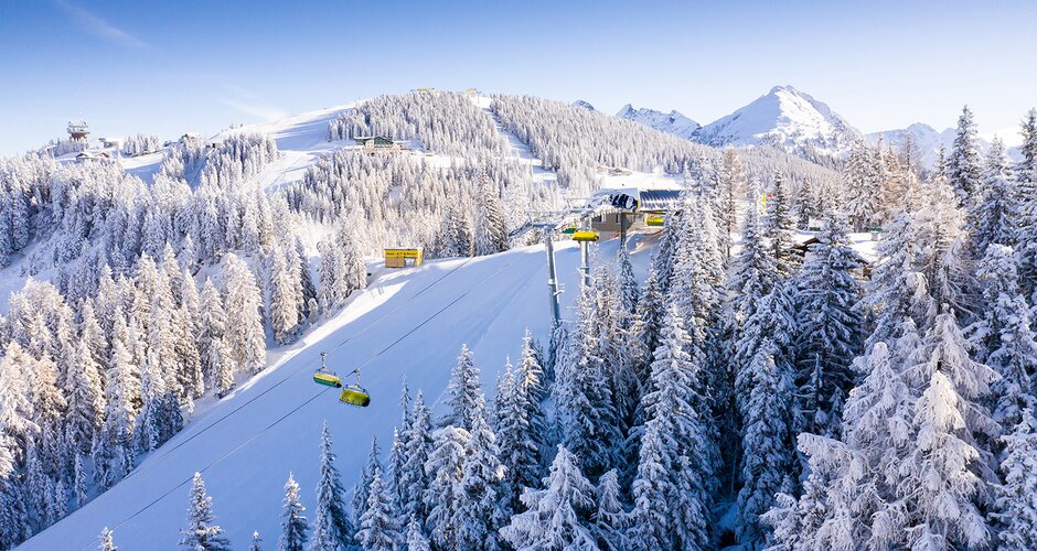 Aerial view of Planai with snowy trees, ski slopes, and a yellow cable car against a blue sky. | © Josh Absenger