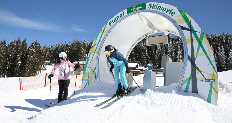 Skier in a blue suit starts through the Skimovie gate on Planai while another skier in pink watches. | © Herbert Raffalt