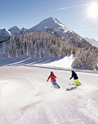 Zwei Skifahrer fahren eine perfekt präparierte Piste vor verschneiter Bergkulisse hinab. | © Herbert Raffalt