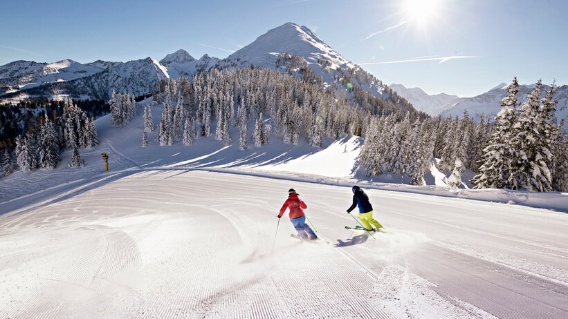 Zwei Skifahrer fahren eine perfekt präparierte Piste vor verschneiter Bergkulisse hinab. | © Herbert Raffalt