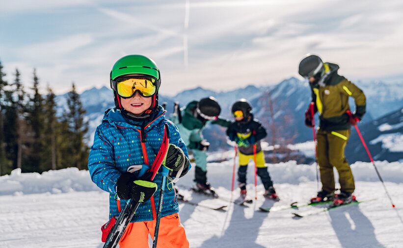 A boy in ski gear stands grinning on the slope and in the background two adults are talking to a child.