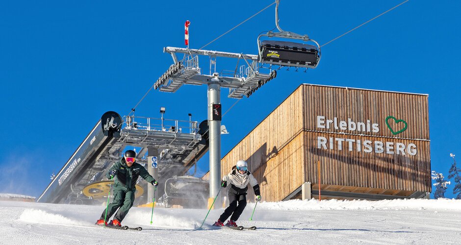 Zwei Skifahrer fahren bei Sonne vor dem Erlebnis Rittisberg und dem Sessellift talwärts.