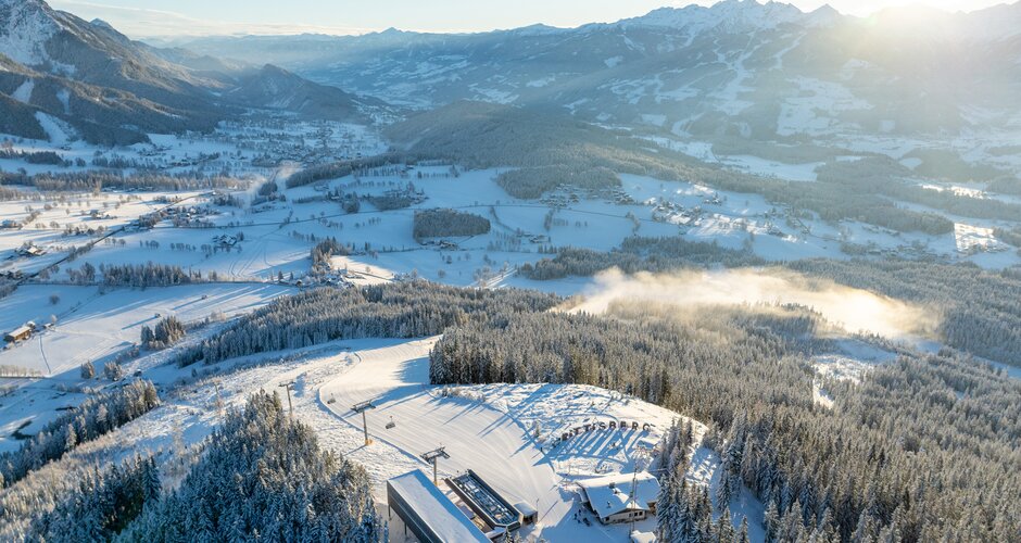 Drohnenaufnahme vom verschneiten Rittisberg mit Talstation, Piste und Ennstal im Sonnenlicht.