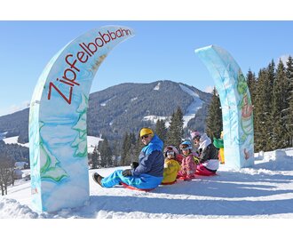Vier Kinder und zwei Erwachsene auf Zipfelbobs unter dem Tor der Zipfelbobbahn | © Hans-Peter Steiner