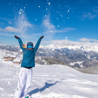 Woman throws snow in the air, smiling, with mountains and snowy valley in background. | © Lorenz Masser