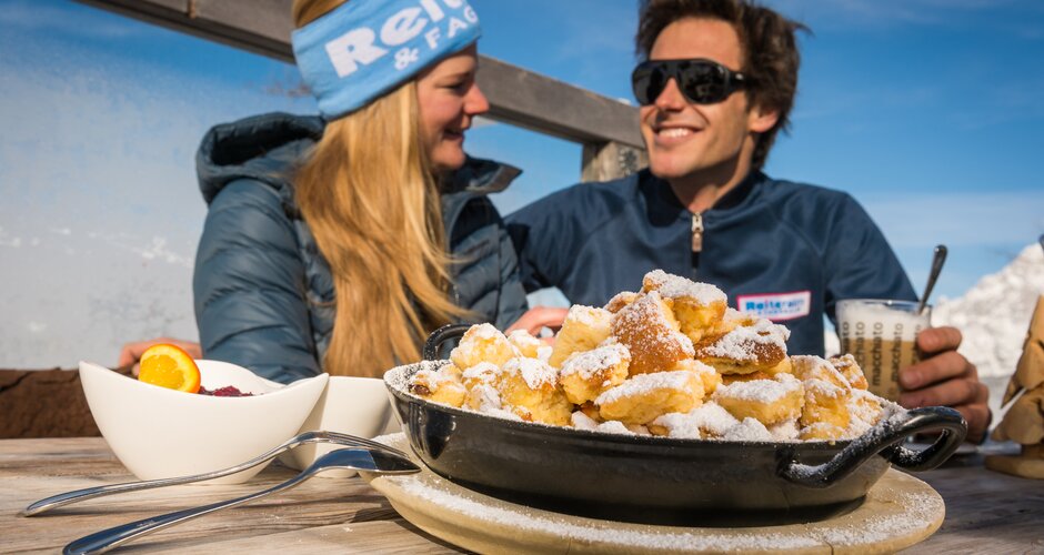 Kaiserschmarrn in a pan on a wooden table, man and woman talking, Reiteralm background. | © Lorenz Masser