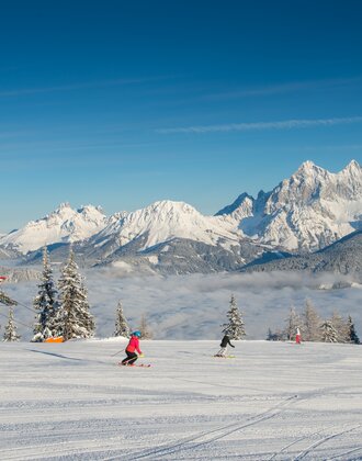 Zwei Skifahrer auf präparierter Piste der Reiteralm, mit verschneitem Dachsteinmassiv im Hintergrund. | © Lorenz Masser
