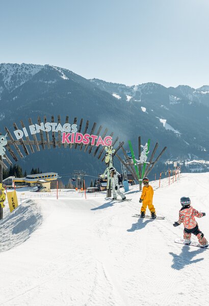Three children in ski or snowboard gear ski towards an arch that says Tuesday Kidsday | © Markus Rohrbacher
