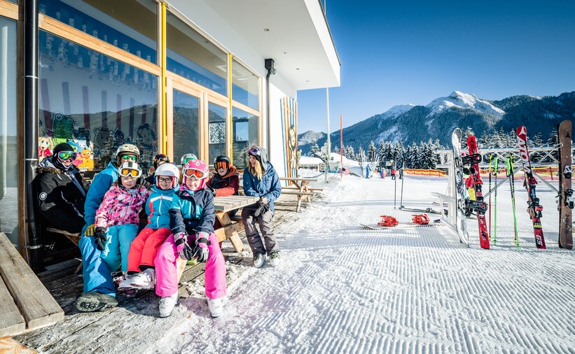 Family in ski clothes sitting on a bench in front of a house and on the opposite side are ski racks | © Markus Rohrbacher