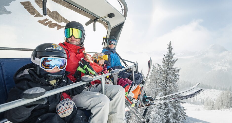 Vierköpfige Familie mit Skihelm und Brille auf Sessellift über verschneiter Winterlandschaft | © Shuttleberg