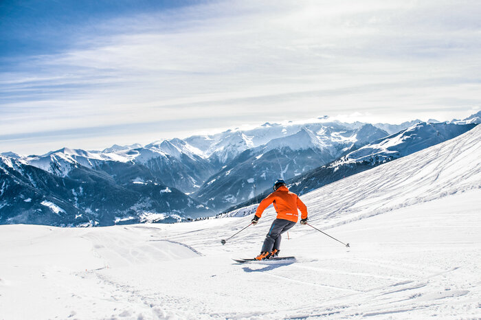 Skier with orange ski jacket descends a groomed slope and snow-capped mountains can be seen in the distance | © Gasteinertal Tourismus GmbH, Creatina