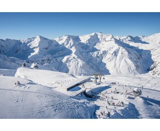 Bergstation einer Kabinenbahn von oben und daneben stehen Skifahrer und rundherum sind schneebedeckte Berge
