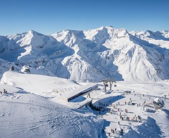 Bergstation einer Kabinenbahn von oben und daneben stehen Skifahrer und rundherum sind schneebedeckte Berge