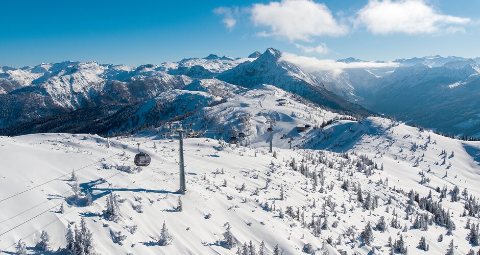 Snow-covered mountain landscape with few trees from above, over which a gondola lift runs and a ski slope can also be seen  | © Snow Space Salzburg