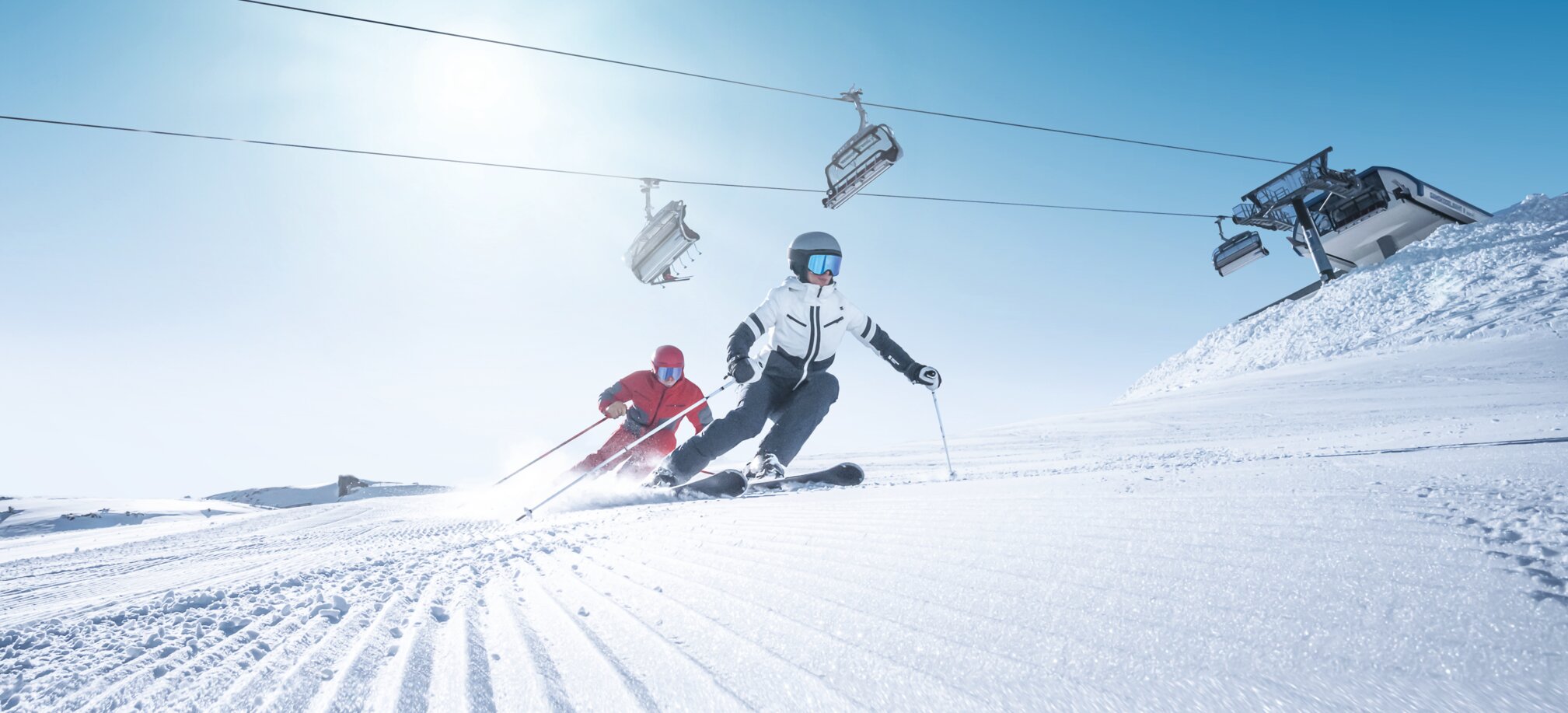 Two skiers carve in sync on freshly groomed slope in sunlight beneath operating chairlift
