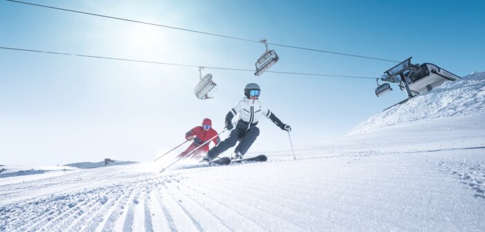 Zwei Skifahrer carven synchron auf frisch präparierter Piste bei Sonnenschein und Liftbetrieb