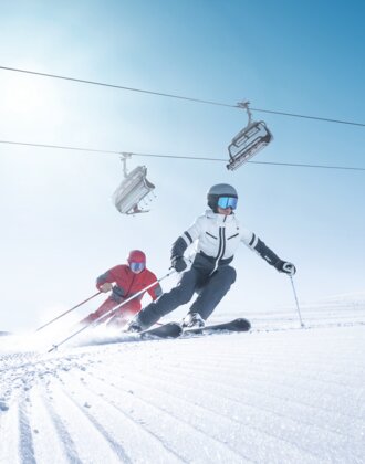Two skiers carve in sync on freshly groomed slope in sunlight beneath operating chairlift