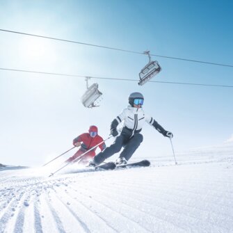 Two skiers carve in sync on freshly groomed slope in sunlight beneath operating chairlift