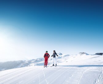 Two skiers stand on a freshly groomed slope with mountain views and sunshine.