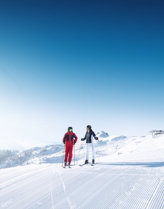 Zwei Skifahrer stehen auf einer frisch präparierten Piste mit Bergpanorama und Sonnenschein.