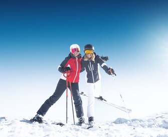 Two female skiers smiling in the snow with poles and helmets in bright sun at Ski amadé