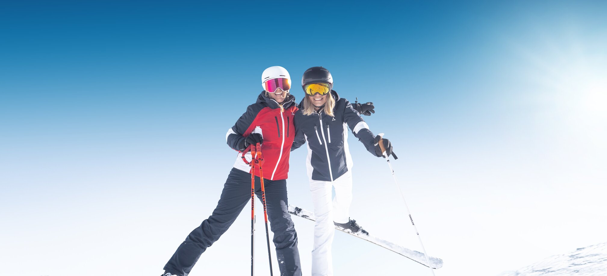 Zwei Skifahrerinnen stehen lachend im Schnee, mit Stöcken und Helmen bei Sonne in Ski amadé