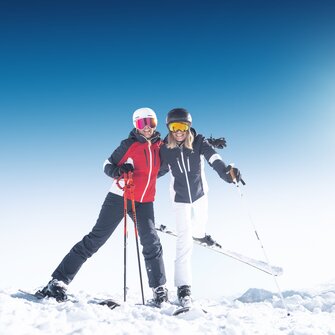 Two female skiers smiling in the snow with poles and helmets in bright sun at Ski amadé