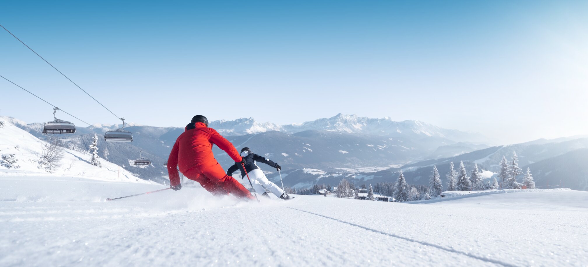 Zwei Skifahrer in rotem und schwarzem Outfit fahren dynamisch eine Piste hinab