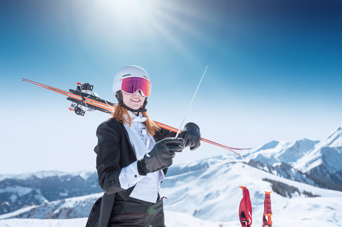 Woman in tuxedo with helmet and goggles holds baton, skis on shoulder, sun shining behind