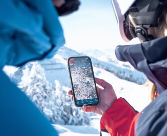 Person wearing ski helmet holds smartphone with slope map open in snowy mountain scenery