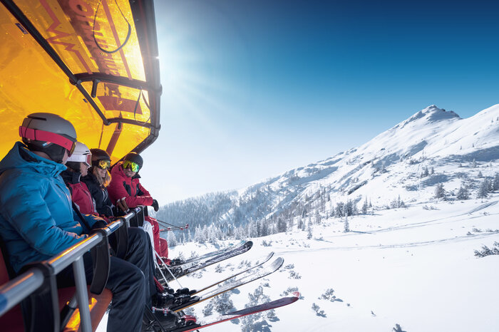 Five skiers sit on a chairlift with orange cover above a snowy mountain landscape
