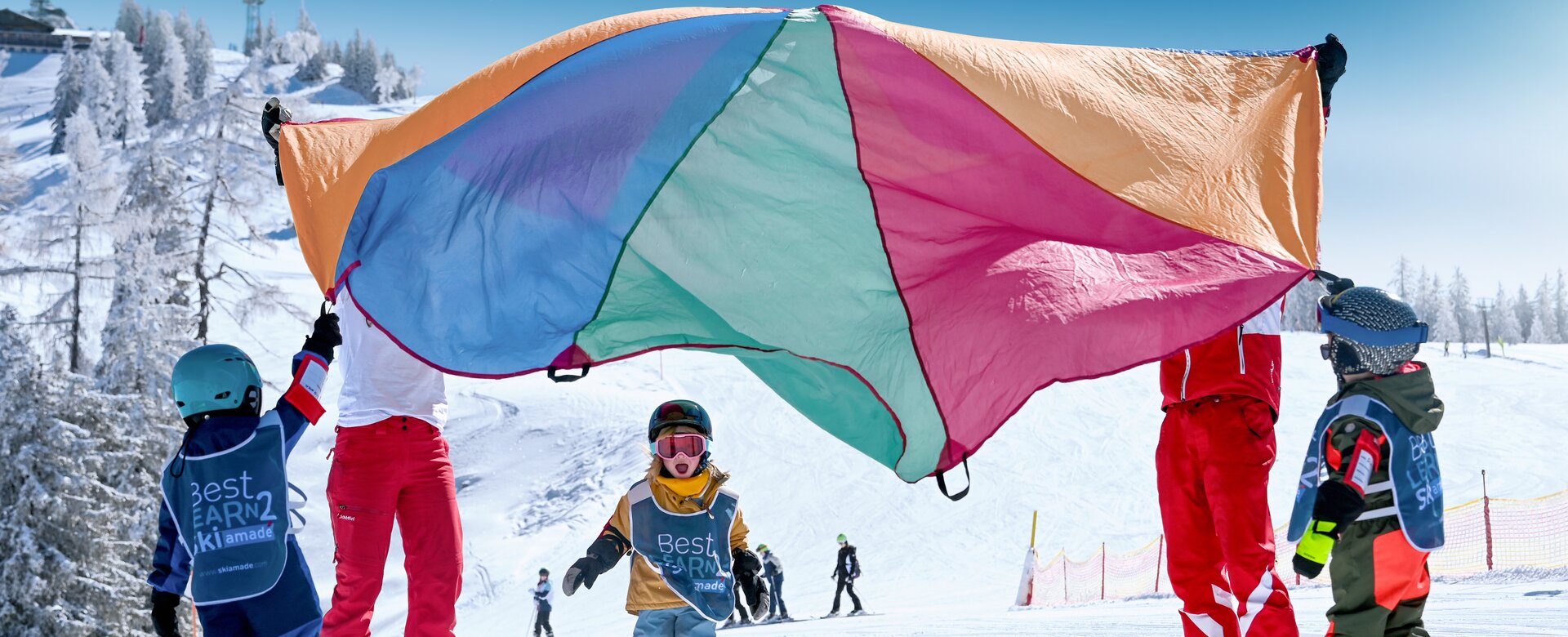 Children in Best Learn2Ski vests play under colourful parachute on snowy slope with ski instructors in the background