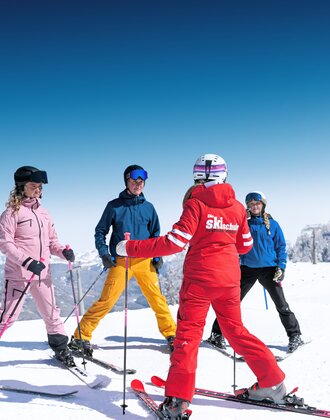 Skilehrerin zeigt vier Skischülern in bunter Kleidung Übungen auf sonniger, verschneiter Piste in Ski amadé