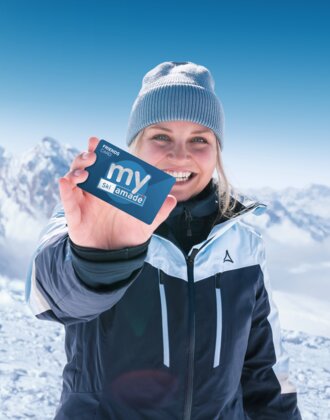 Frau in Skijacke hält my Ski amadé Friends Card vor schneebedeckten Alpen bei strahlendem Sonnenschein