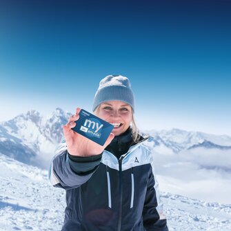 Frau in Skijacke hält my Ski amadé Friends Card vor schneebedeckten Alpen bei strahlendem Sonnenschein