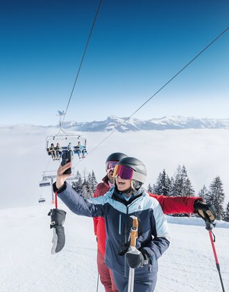 Zwei Skifahrer machen ein Selfie auf sonniger Piste mit Sessellift und Alpenblick in Ski amadé