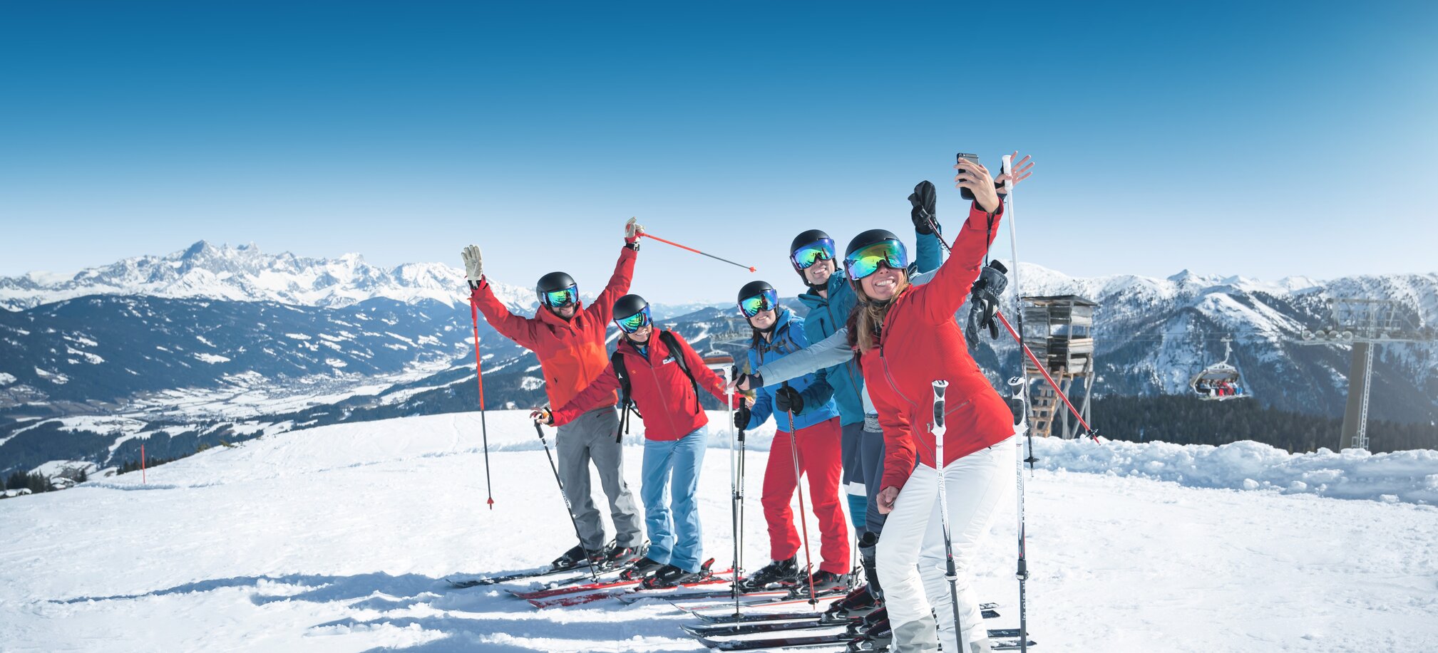 Six skiers take a cheerful group selfie on the slope in sunny weather with mountains behind