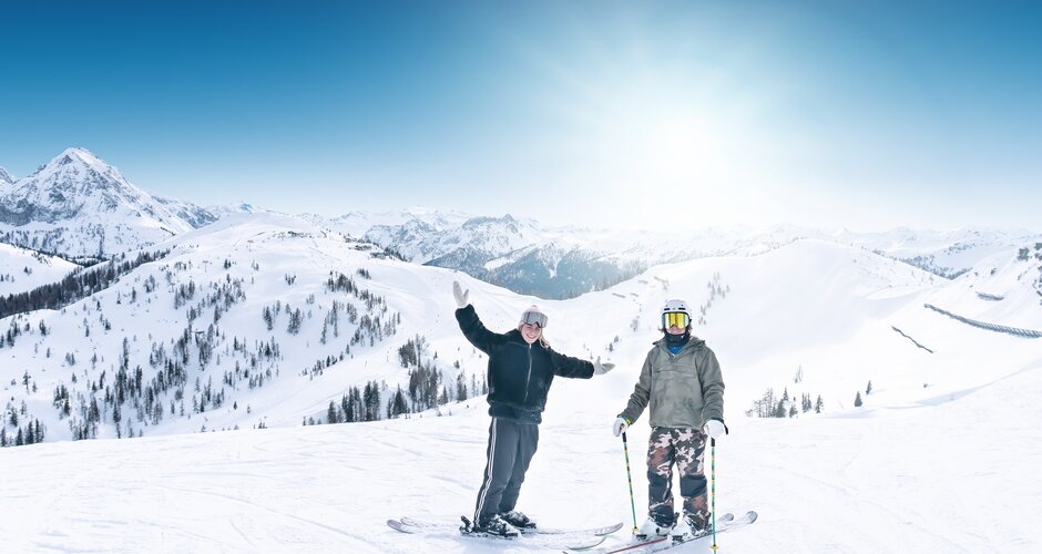 Two skiers stand on a sunny slope with a wide view of snow-covered mountain landscape