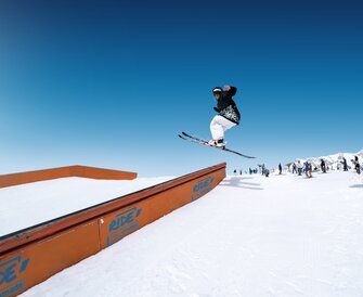 Skier jumps over rail element in snow park, other winter sports fans watching in the background