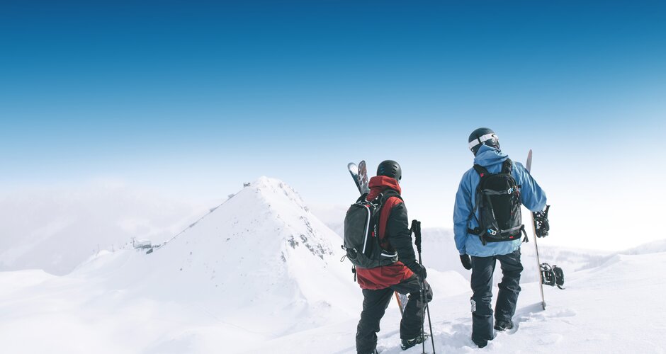 Two winter sports athletes with skis and snowboard stand on snowy slope facing mountain peaks