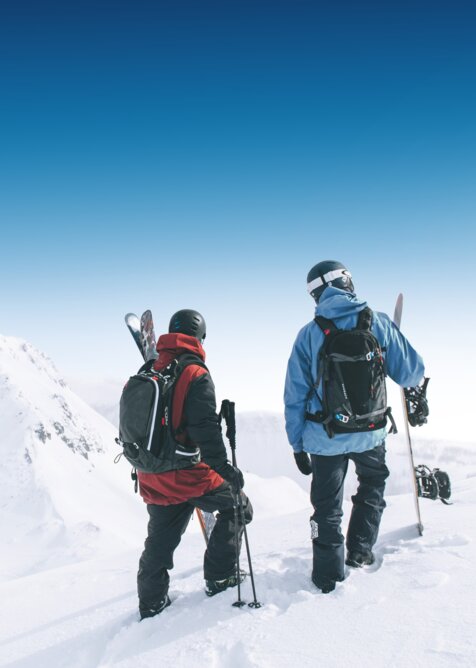 Two winter sports athletes with skis and snowboard stand on snowy slope facing mountain peaks