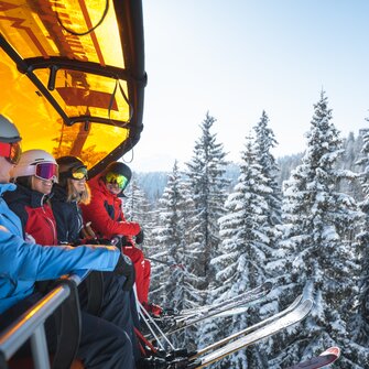 Vier Skifahrer mit Helmen und Skibrillen sitzen auf einem Sessellift mit orangener Haube über verschneiten Tannen. | © Ski amadé