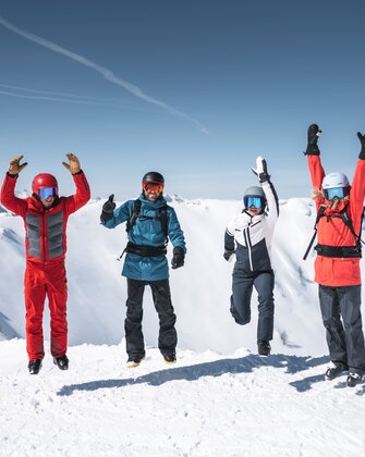 Vier Skifahrer springen gleichzeitig in die Luft, Schneeberge und Sonne im Hintergrund