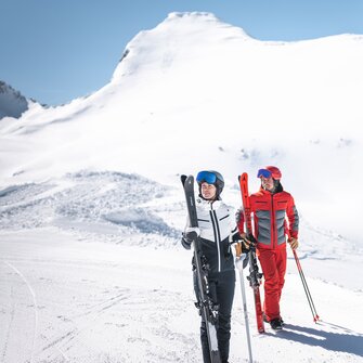 Two skiers with skis and poles stand on groomed slope, with snowy mountain scenery in the background.